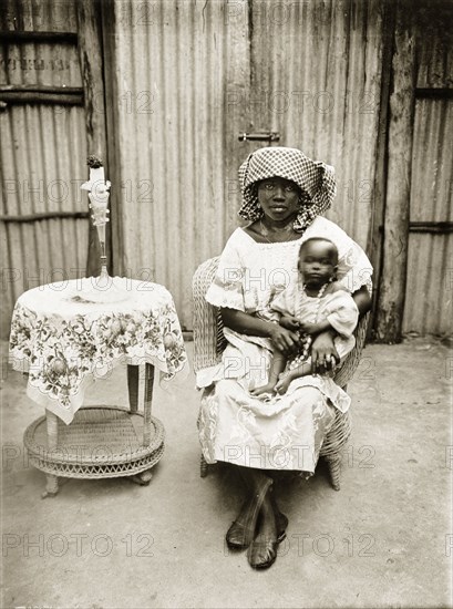 Portrait of a Nigerian woman and baby. The woman and baby are seated in a cane chair in front of a corrugated iron wall and gates. Next to them is a small cane table with a floral cloth and vase. Badagry, Nigeria, circa 1928. Badagry, Lagos, Nigeria, Western Africa, Africa.