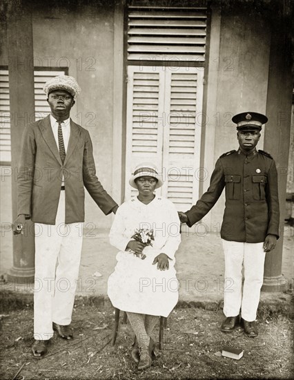 Portrait of Salvation Army workers. Two African men in Western dress stand on either side of a seated woman holding a bunch of flowers. The man on the right wears a Salvation Army cap and jacket, the woman wears a long-sleeved dress, thick tights, and high-heeled leather shoes. She also wears a Salvation Army hat. Badagry, Nigeria, circa 1928. Badagry, Lagos, Nigeria, Western Africa, Africa.