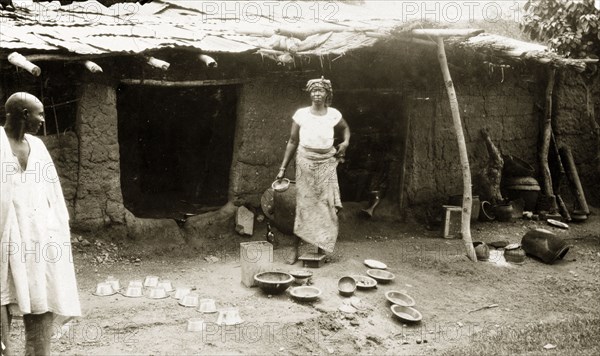 Selling brass pots, Nigeria. A woman with brass pots to sell outside her mud house. Nigeria, circa 1925. Nigeria, Western Africa, Africa.