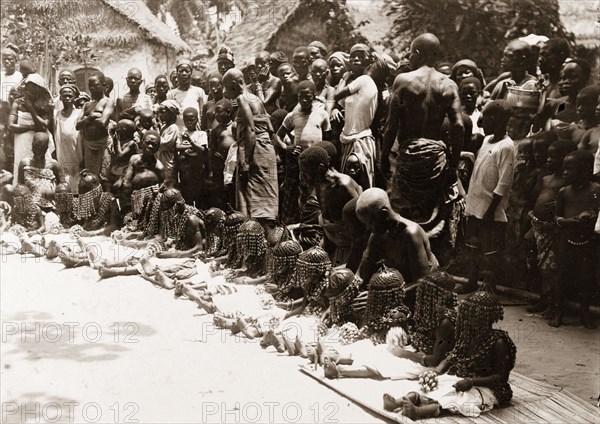 Children's festival, Nigeria. A line of young children wait to perform a traditional dance at a children's festival. Seated on mats, they wear caps of leather or gourd with veils made from beaded shells hiding their faces. A crowd of older children and adults stands behind them. Nigeria, circa 1925. Nigeria, Western Africa, Africa.