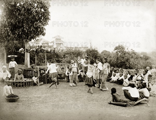 Children perform a 'pwe'. Several young girls perform a 'pwe' (Burmese dance drama) surrounded by a small audience and supported by two traditional Burmese drummers and other musicians. Masks hang from a framework suspended above one of the circular drum units. Burma (Myanmar) circa 1900. Burma (Myanmar), South East Asia, Asia.