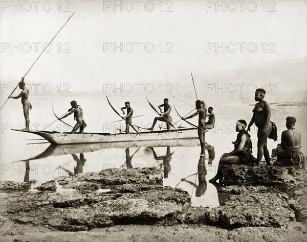 Andaman Islanders fishing. Six nearly naked men stand in a canoe, or in shallow water beside it, poised with bows and arrows ready to catch fish. Three women sit or stand on rocks at the water's edge. Andaman Islands, India, circa 1900. India, Southern Asia, Asia.