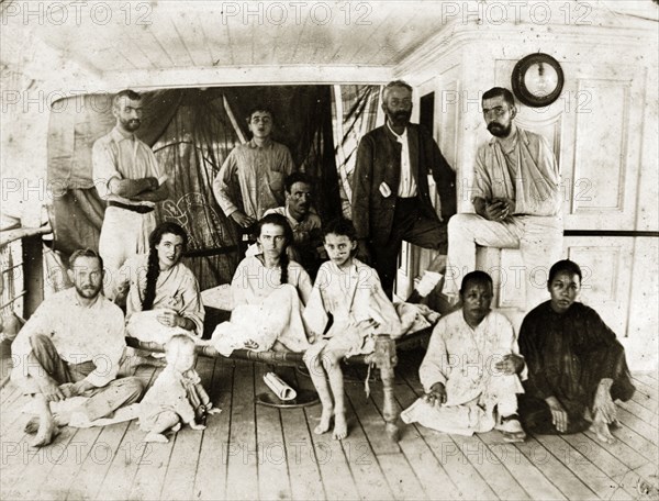 Survivors from SS Aden. Twelve men, women and children in informal dress pose on the deck of a ship following their rescue from the SS Aden. Two of the European women and a child are seated on an Indian string cot: two Asian women squat on the floor. Indian Ocean near Socotra, June 1897. Socotra, Adan, Yemen, Middle East, Asia.