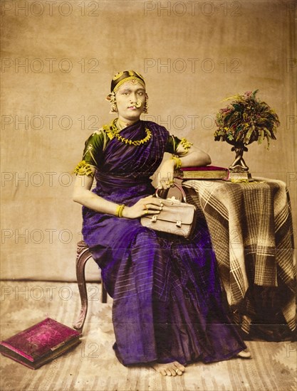 Southern Indian woman. Portrait of a southern Indian woman seated on a European-style chair beside a small covered table with a vase. She wears a heavy sari over a short-sleeved blouse, adorned with gold jewellery, including bracelets, upper arm pieces, earrings and ear tops, nose ring and necklace. She holds a leather handbag and her feet are bare, save for a toe ring. India, circa 1885. India, Southern Asia, Asia.
