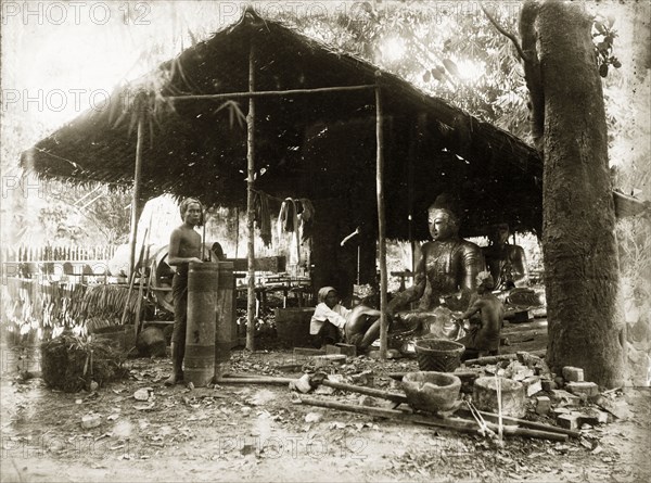 Idol-making, Burma (Myanmar). Two craftsmen sit, under a thatched, open-walled shelter, burnishing or painting a large cross-legged statue of the Buddha. A woman squats beside them. A second statue is visible in the background: a collection of metalworking tools in foreground. Burma (Myanmar), circa 1900. Burma (Myanmar), South East Asia, Asia.
