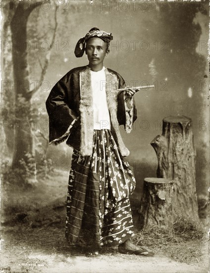 Portrait of a Burmese gentleman. A posed studio portrait of a finely-dressed Burmese gentleman. His outfit comprises a fur-lined silk jacket, headscarf and a traditional silk 'longyi' or wraparound skirt. He poses for the camera holding a smoking cheroot (a type of cigar). Burma (Myanmar), circa 1885. Burma (Myanmar), South East Asia, Asia.