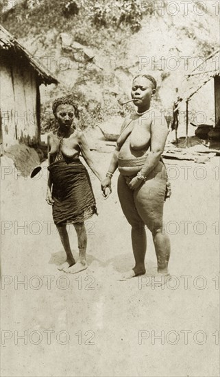 Two women of the Andaman Islands. Two standing women hold hands against a backdrop of a cliff and makeshift huts. One wears a cloth wrapped loosely around her waist. The other who is naked apart from a belt, necklace and bracelets, smokes a pipe. Andaman Islands, India, circa 1900., Andaman and Nicobar Islands, India, Southern Asia, Asia.