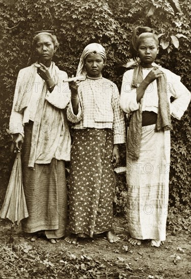 Three Burmese women. Outdoors portrait of three young Burmese women wearing traditional dress. Their hair is worn up and two of the girls smoke cheroots (Burmese cigars). Burma (Myanmar), circa 1900. Burma (Myanmar), South East Asia, Asia.