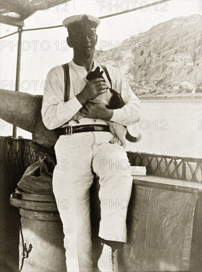 Gunner aboard 'Karanja'. Mr Harknet', a gunner aboard the armed launch boat 'Karanja', holds a domestic cat to his chest as he poses on deck. Probably the Persian Gulf, circa 1900., Middle East, Asia.