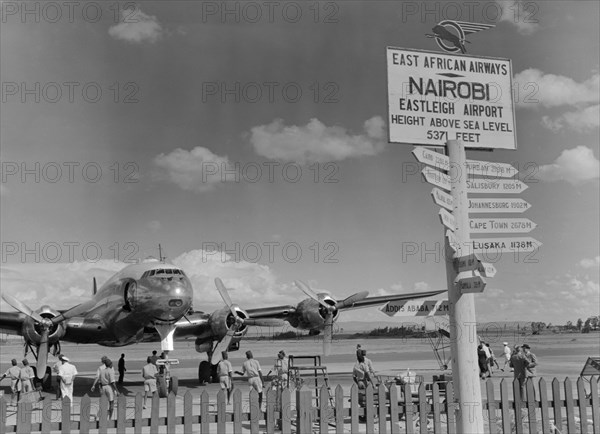 Eastleigh Airport, Nairobi. Airport staff mill around a double-propellered passenger plane that sits on the runway at Eastleigh airport. A sign post on the right indicates the distance in miles to various African cities. Nairobi, Kenya, 9 November 1952. Nairobi, Nairobi Area, Kenya, Eastern Africa, Africa.