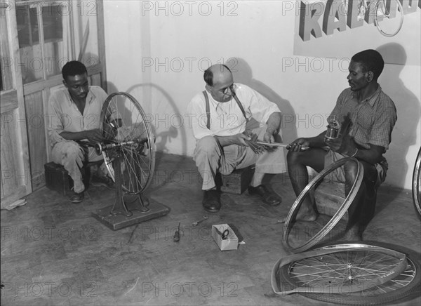 Kassam Kanji's workshop'. Two Kenyan men work alongside a European, repairing bicycle wheels in a workshop identified as 'Kassam Kanji's'. Kenya, 22 November 1952. Kenya, Eastern Africa, Africa.