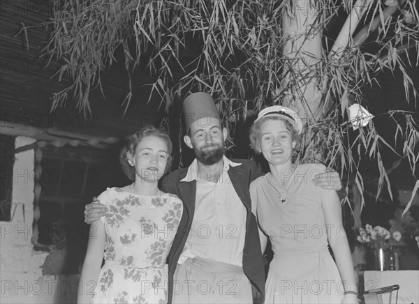 Charles Trotter at a Christmas party. Charles Trotter, a professional photographer whose work is widely represented in this collection, enjoys himself at a Christmas party. He sports a fake fez hat and poses with two women identified as 'Barbara and Mary' outside the Forest Inn bar. Possibly Nairobi, Kenya, 25 December 1952. Kenya, Eastern Africa, Africa.
