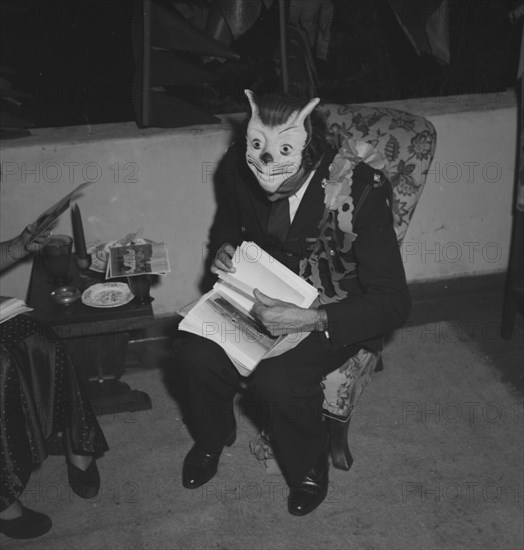 Ron Richardson with mask. Ron Richardson takes a break from the merriment at the Archer's New Year's party. Decorated with a paper garland, he sits engrossed in a set of papers, his fancy dress mask pushed up on top of his head. Kenya, 31 December 1952. Kenya, Eastern Africa, Africa.