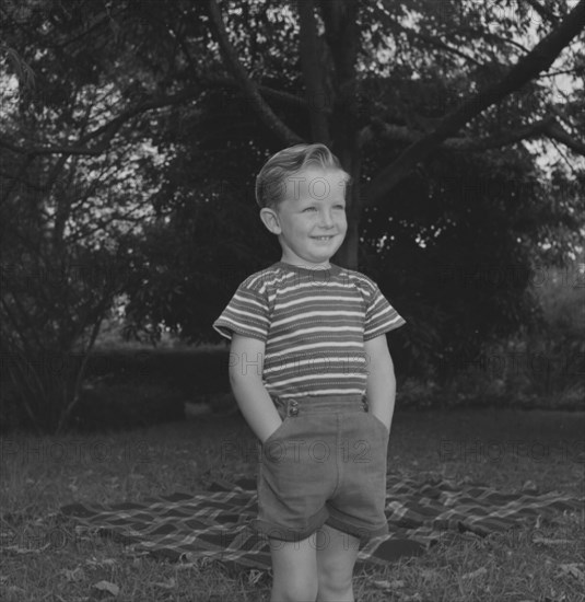 Cheerful boy, Kenya. Stephen Ward stands, hands in pockets, smiling broadly beside a picnic rug laid out in a garden. Kenya, 1 January 1953. Kenya, Eastern Africa, Africa.