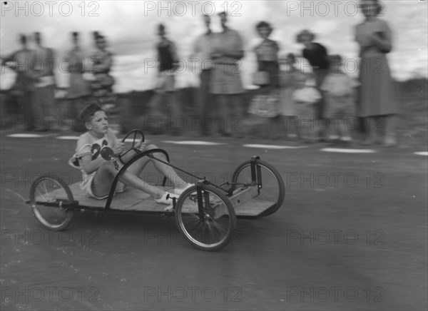 Number 26 in the soap box derby. A young boy braces himself as he whizzes past crowds riding a homemade go-cart, entry number 26 in a soap box derby race. Kenya, East Africa, 03 January 1953. Kenya, Eastern Africa, Africa.