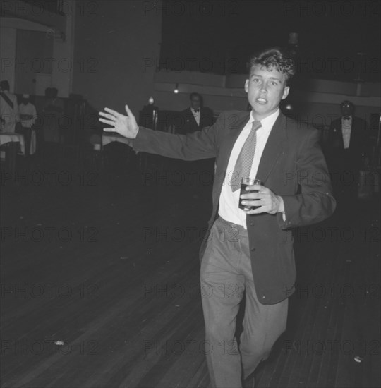 On the dance floor. A young man dressed in a suit approaches the camera, glass in hand, from across an empty dance floor at a Jazz Ball. Kenya, 21 September 1957. Kenya, Eastern Africa, Africa.