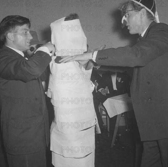 The Mogambo mummy. Members of a rugby team dressed in tuxedos fool around at the Mogambo Club. One of the men has been 'mummified' in toilet paper by his friends, one of whom wears a false nose and moustache. Possibly Nairobi, Kenya, 21 September 1957. Kenya, Eastern Africa, Africa.