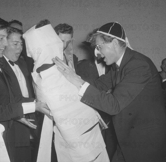 The Mogambo mummy. Members of a rugby team dressed in tuxedos fool around at the Mogambo Club. One of the men has been 'mummified' in toilet paper by his friends, one of whom wears a false nose and moustache. Possibly Nairobi, Kenya, 21 September 1957. Kenya, Eastern Africa, Africa.