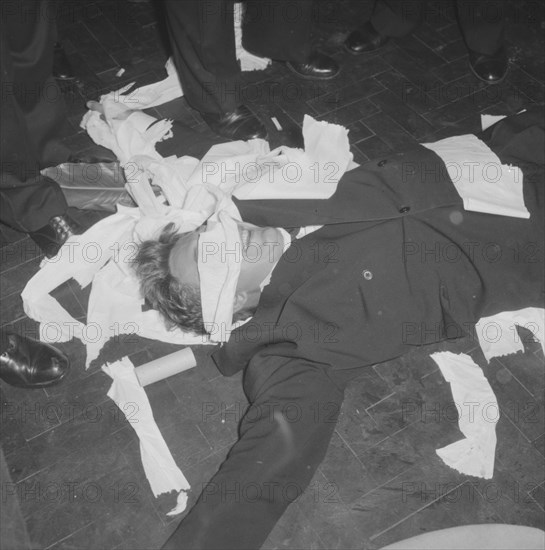 Rugby team antics. A victim of rugby team antics at the Mogambo Club lies spread-eagled on the floor. He has just de-tangled himself after having been wrapped in toilet paper by his friends. Possibly Nairobi, Kenya, 21 September 1957. Kenya, Eastern Africa, Africa.