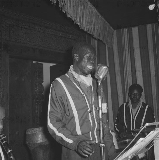 Singer at the Equator Club. An African man sings into a microphone on a small stage at the Equator Club. Other members of the band can be seen behind him, playing instruments including drums and a clarinet. Nairobi, Kenya, 21 September 1957. Nairobi, Nairobi Area, Kenya, Eastern Africa, Africa.