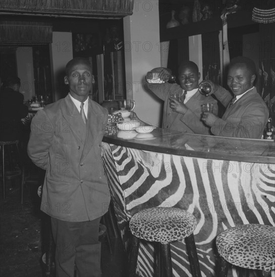 Bartenders at the Equator Club. Portrait of three uniformed waiters at the Equator Club. Two African bartenders jokingly pour drinks into glasses behind a bar decorated with zebra stripes and leopard-print stools. Nairobi, Kenya, 21 September 1957. Nairobi, Nairobi Area, Kenya, Eastern Africa, Africa.