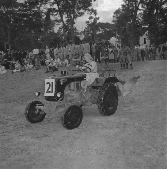 Tractor in a vehicle parade. A young woman with coiffeured hair and heels, drives a small tractor past a crowd of onlookers in a vehicle parade at the Royal Show. Kenya, 26 September 1957. Kenya, Eastern Africa, Africa.