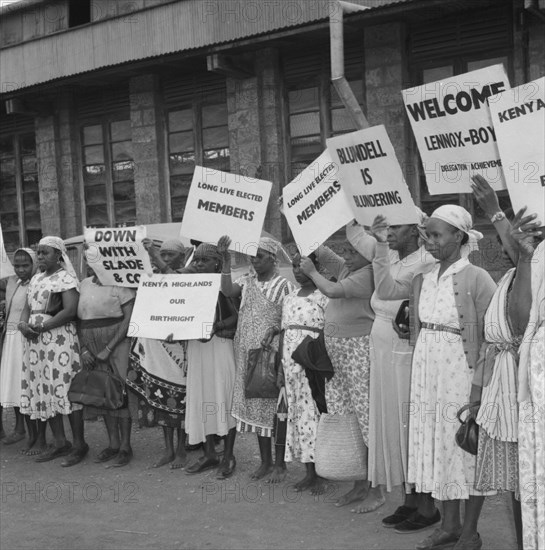 Female Kenyan demonstrators. Kenyan women involved in a peaceful demonstration at an airport, hold up placards on the arrival of politican Alan Tindal Lennox-Boyd. Amongst these are two that read: 'Blundell is Blundering' and 'Long Live Elected Members'. Kenya, 11 October 1957. Kenya, Eastern Africa, Africa.