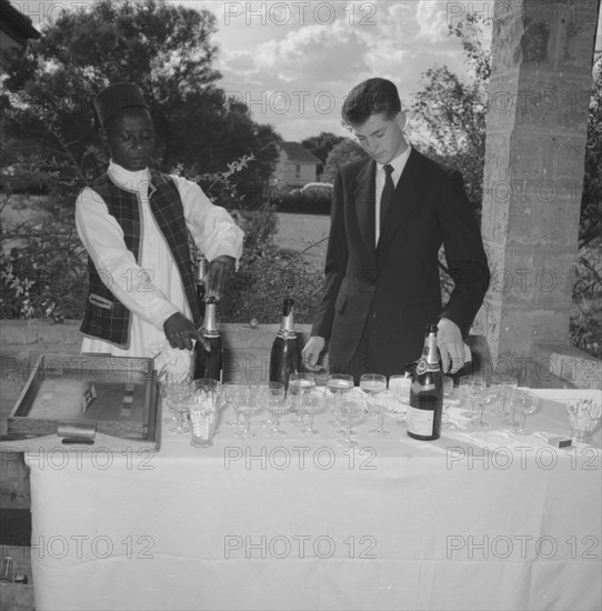 Wedding barmen. A young European man assists a uniformed African servant to pour champagne at the Hodgson-Wilkinson wedding. Kenya, 12 October 1957. Kenya, Eastern Africa, Africa.