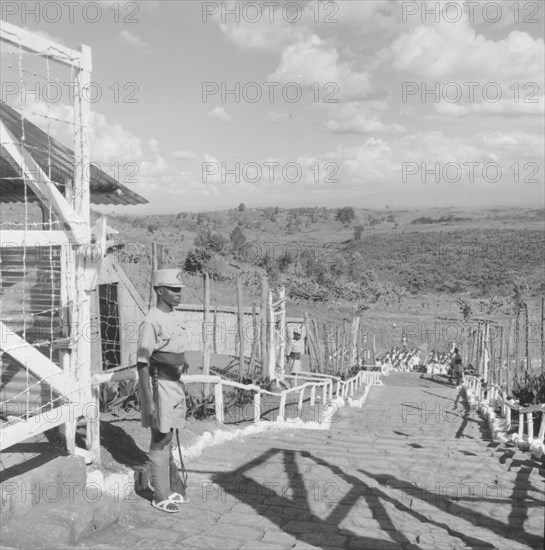 Fort Hall military camp. Armed police guard the steps of a fenced military camp for Kenyan detainees. This photograph was taken during Lennox-Boyd's visit to the Fort Hall district. Kangema, Kenya, 15 October 1957. Kangema, Central (Kenya), Kenya, Eastern Africa, Africa.