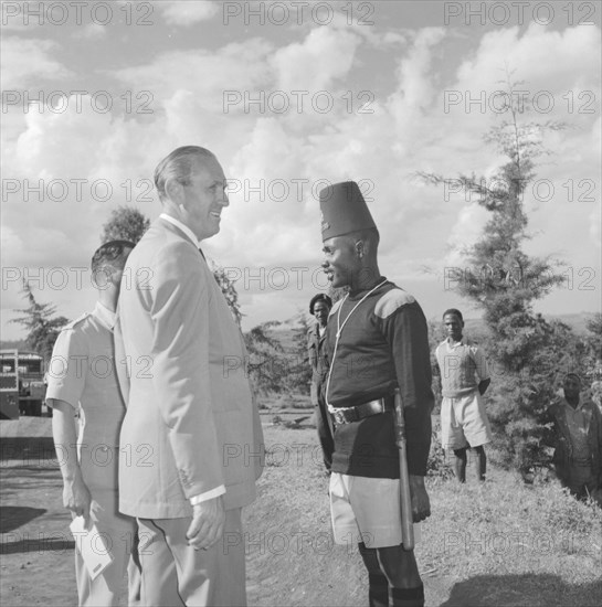 Lennox-Boyd visits Fort Hall. Alan Tindal Lennox-Boyd (1904-1983) stands on a roadside beside a uniformed askari (soldier) on his visit to the Fort Hall district. Kangema, Kenya, 15 Oct 1957. Kangema, Central (Kenya), Kenya, Eastern Africa, Africa.