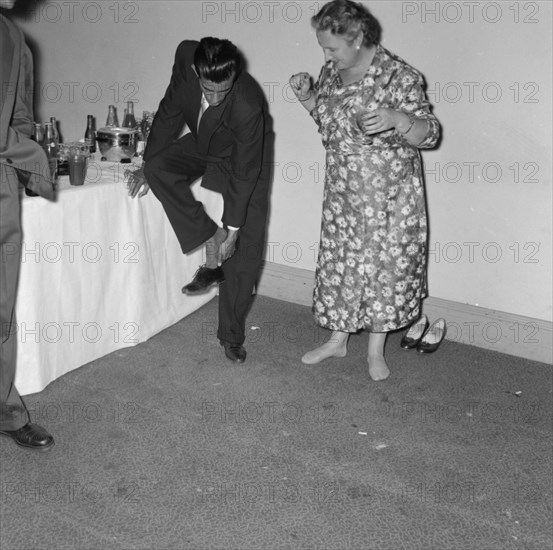 Removing shoes at a cocktail party. A suited man leans on a table for support as he removes his shoe at a Prudential Insurance cocktail party. He is encouraged by a middle-aged woman who is already barefoot and ready to dance. Kenya, 16 October 1957. Kenya, Eastern Africa, Africa.