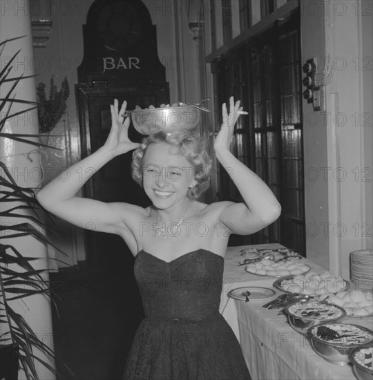 A well balanced diet. A woman dressed in an evening gown and jewellery attempts to balance a bowl of food on her head at the Limuru Hunt Ball buffet. Kenya, 19 October 1957. Kenya, Eastern Africa, Africa.
