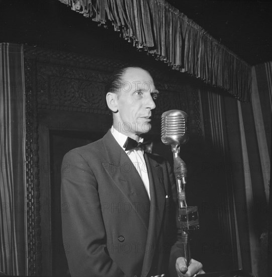 On stage at the Equator Club. A European man stands before a microphone on a small, curtained stage at the Equator Club. Related images suggest he is a singer with a band of African musicians. Nairobi, Kenya, 19 October 1957. Nairobi, Nairobi Area, Kenya, Eastern Africa, Africa.