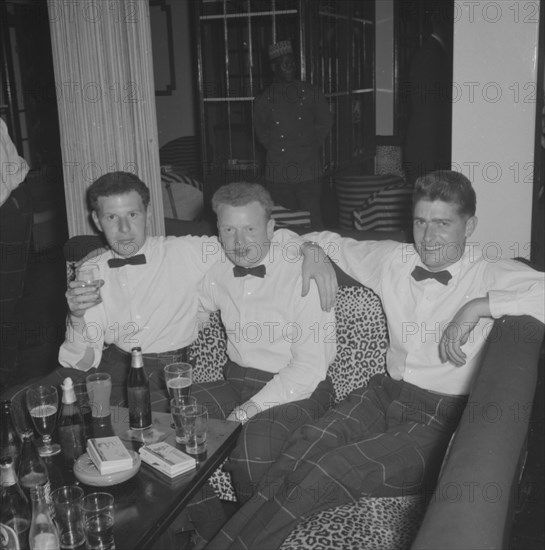 Three Cameronians. Three young Cameronians enjoy themselves at the Equator Club. Dressed in matching check trousers and bow ties, they sit around a table covered with drinks on a leopard print sofa. Nairobi, Kenya, 19 October 1957. Nairobi, Nairobi Area, Kenya, Eastern Africa, Africa.
