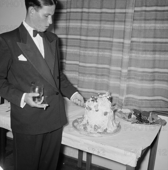 Cutting the cake. A young man dressed in a tuxedo cuts his 21st birthday cake, drink and cigarette in hand, at the Orme-Smith party. Kenya, 9 November 1957. Kenya, Eastern Africa, Africa.