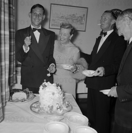 Cutting the cake. A young man dressed in a tuxedo, holds up a knife for the camera before cutting into his 21st birthday cake at the Orme-Smith party. He is surrounded by middle-aged well-wishers, perhaps family members, who stand beside him, bowls at the ready. Kenya, 9 November 1957. Kenya, Eastern Africa, Africa.