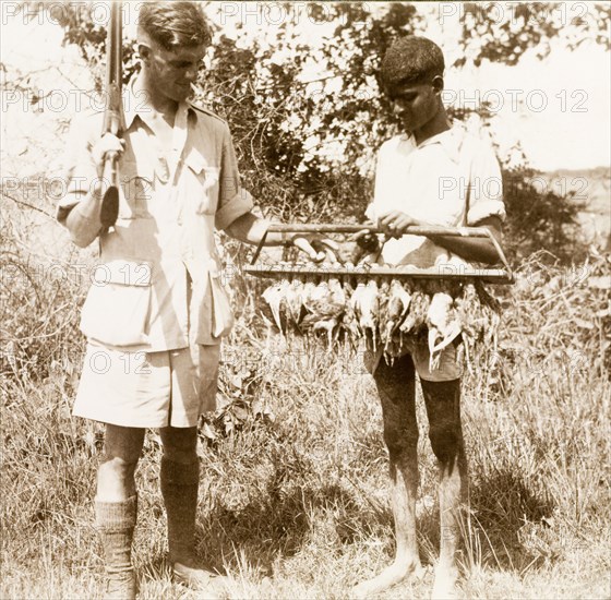 Bird shooting at Gokalpur. A British man identified as 'D.R.G.' ('Dennis') smokes a cigarette, shotgun over shoulder, as he examines the day's kill: a rack full of birds carried by an Indian servant. Gokalpur, India, circa 1943. Gokalpur, West Bengal, India, Southern Asia, Asia.