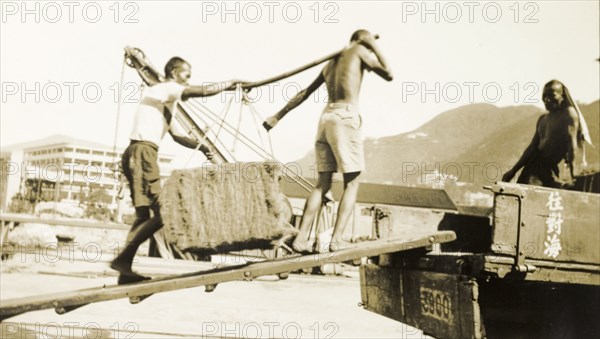 Asian workers, Kowloon. Asian workers carry a heavy load up a ramp onto the back of an open truck. The cargo is unidentified, but an original caption comments that it is heavy, weighing in at 260 pounds (118 kilograms). Kowloon, Hong Kong, (People's Republic of China), 1937. Kowloon, Hong Kong, China, People's Republic of, Eastern Asia, Asia.