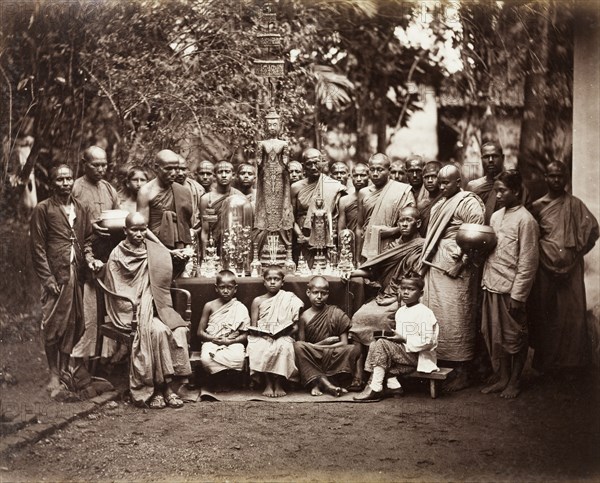 Buddhist monks with religious icons. A group of Buddhist monks stand around a table laden with religious icons including metal statues, figurines and vessels. Two robed monks are seated on either side of the table whilst four young boys squat on stools at the front of the group. Ceylon (Sri Lanka), circa 1875. Kandy, Central (Sri Lanka), Sri Lanka, Southern Asia, Asia.