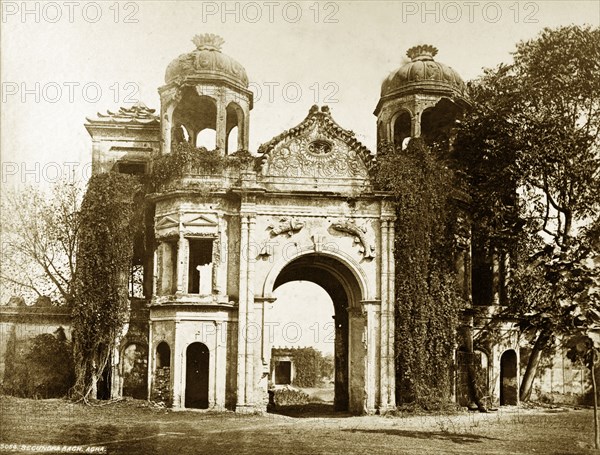 Gateway to the Sikanderbagh. An ornate gateway, partially covered by vegetation, features the fish or 'macchi' that were a traditional symbol of the nawabs of Awadh. The ruins of the Sikanderbagh Palace can be glimpsed through the arch. Lucknow, North Western Provinces (Uttar Pradesh), India, circa 1875. Lucknow, Uttar Pradesh, India, Southern Asia, Asia.