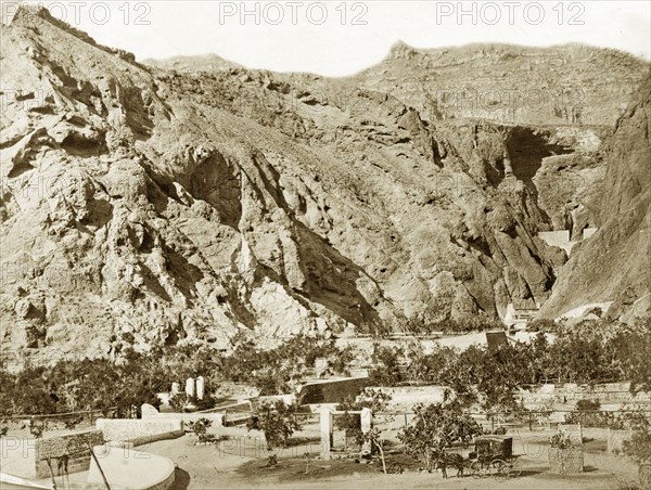 Water wells, Aden. Steep mountains rise up immediately behind several masonry wells, which are sited on a plain dotted with trees. A horse and carriage stands by one of the wells. Aden, Yemen, circa 1885. Aden, Adan, Yemen, Middle East, Asia.