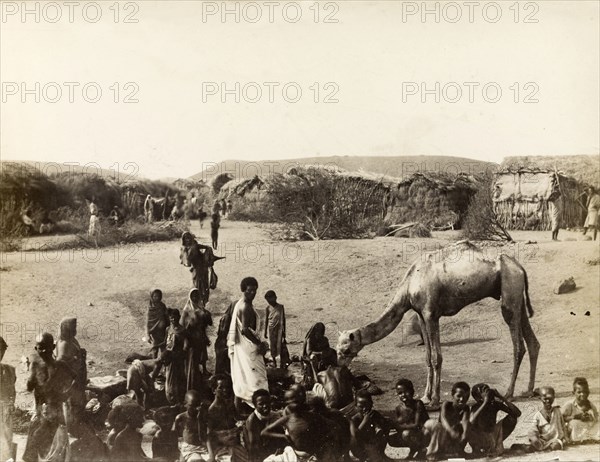 Village scene at Aden. A cluster of people, many of them children, stand or squat on the sand in front of a village of grass huts. A camel is pictured with the group. Aden, Yemen, circa 1885. Aden, Adan, Yemen, Middle East, Asia.