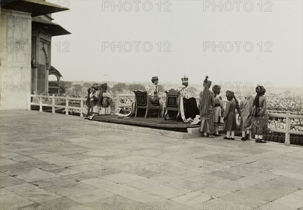 The royal couple at the Delhi Fort. King George V (r.1910-36) and Queen Mary appear before crowds from a throned balcony at the Delhi Fort. Pictured the day after the Coronation Durbar, they are dressed in their coronation robes and crowns, and are accompanied by ten Indian pages. Delhi, India, 13 December 1911. Delhi, Delhi, India, Southern Asia, Asia.