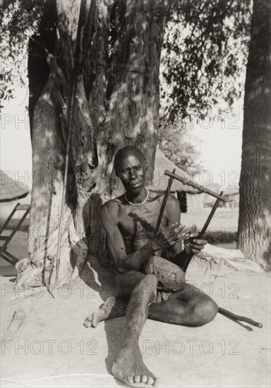 A Zande hand harp. A Zande man plays a harp as he sits beneath a tree. The photographer comments: "The Zande people enjoyed music, and you might meet them on a bush path playing a sort of hand harp made of vibrating metal strips attached to a small wooden block". Meridi, Sudan, circa 1947. Meridi, Upper Nile, Sudan, Eastern Africa, Africa.