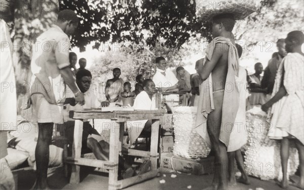 Cotton marketing at Meridi. Traders queue at an open-air market, balancing large, open-weave baskets of seed cotton on their heads, ready to be graded, weighed and sold. The photographer comments: "The weighing machine (was) prepared and people would line up according to their grade, have their load weighed, and then present their ticket for payment". Meridi, Sudan, 1947. Meridi, Upper Nile, Sudan, Eastern Africa, Africa.