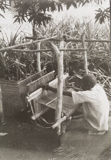 A Zande weaver at work. A Zande weaver sits outdoors on a rush mat as he operates a hand loom. Near Meridi, Upper Nile State, Sudan, circa 1947. Meridi, Upper Nile, Sudan, Eastern Africa, Africa.