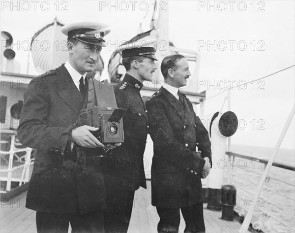 Photography from the S.S. Balmoral Castle. Two British naval officers admire the view from the deck of S.S. Balmoral Castle, whilst another prepares to take a photograph. The ship was transporting the Duke and Duchess of Connaught to South Africa, where they were due to open the new Union Parliament in Cape Town. Probably Atlantic Ocean, Africa, October 1910. Africa.