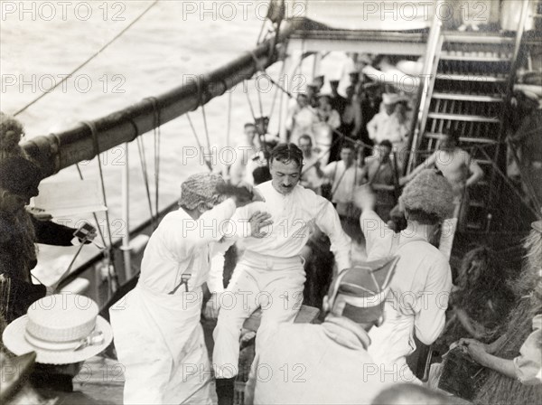 Antics aboard the S.S. Balmoral Castle. Crew members aboard the S.S. Balmoral Castle clown about on deck wearing costumes and wigs, possibly for a traditional 'crossing the line' naval initiation ceremony. The ship was transporting the Duke and Duchess of Connaught to South Africa, where they were due to open the new Union Parliament in Cape Town. Probably Atlantic Ocean, Africa, October 1910. Africa.