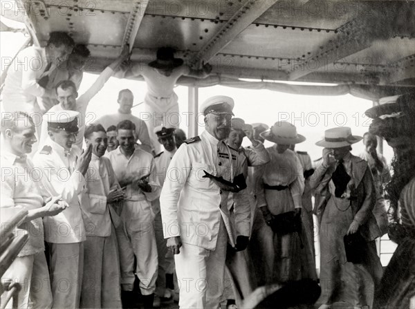 The Duke of Connaught joins in the fun. The Duke of Connaught is pictured with a fish tied around his neck as he participates in antics aboard the S.S. Balmoral, watched by a crowd of crew members and his wife, who are clearly entertained. Probably Atlantic Ocean, Africa, October 1910. Africa.