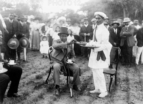 Tea and cake at a royal reception. Dressed in a Western-style suit and top hat, an African chief (possibly Litunga Lubosi I of Barotseland, r.1885-1916) accepts a cup of tea and a slice of cake from an Indian servant at an outdoor reception for the Duke and Duchess of Connaught. This was one of several stops made by the royal couple following their official visit to Cape Town to open the new Union Parliament. Livingstone, Northern Rhodesia (Zambia), 12 November 1910. Livingstone, South (Zambia), Zambia, Southern Africa, Africa.